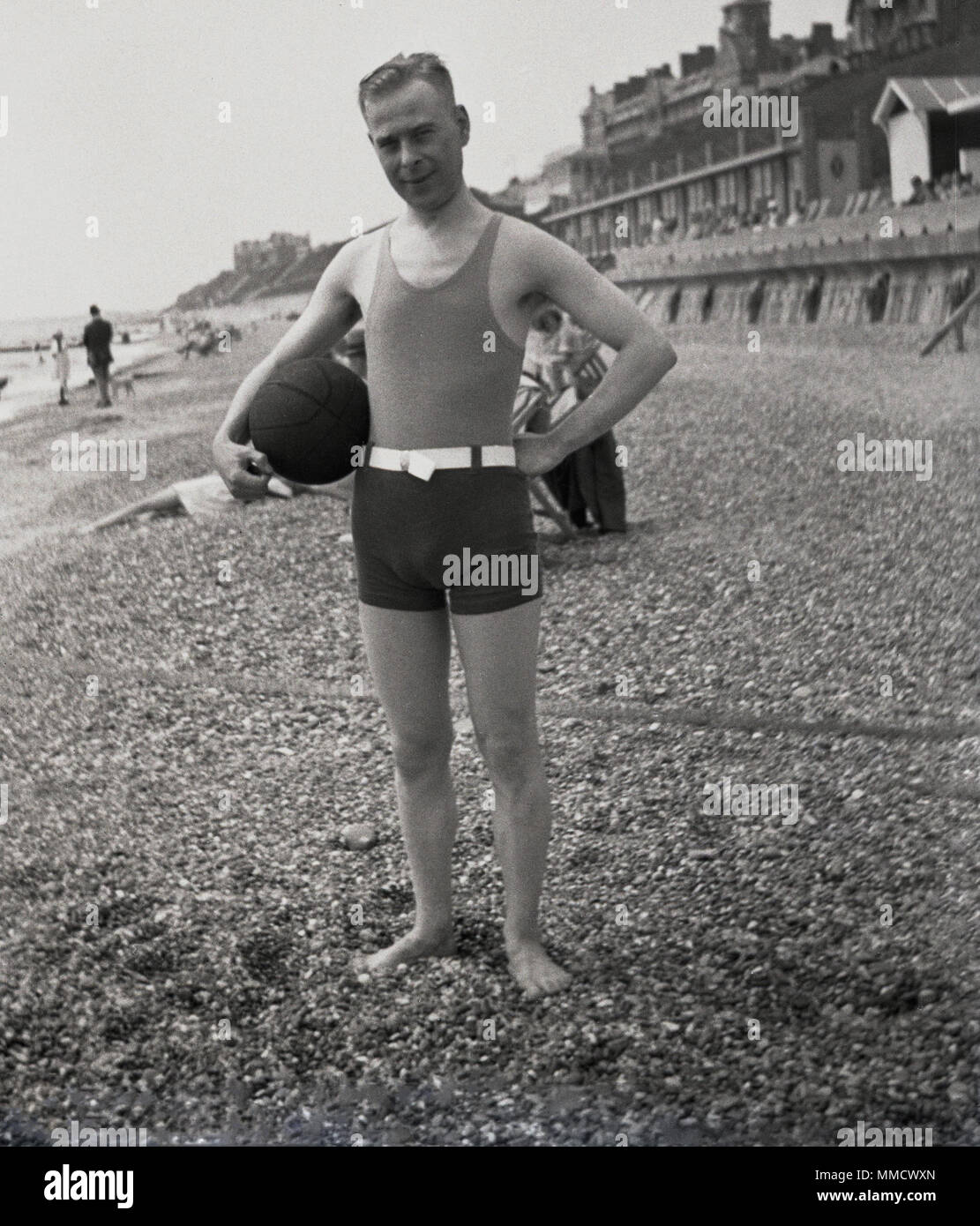 1930s, uomo adulto indossando il costume da bagno dell'epoca, un elegante  pezzo gilet e shorts, in piedi su una spiaggia di ciottoli beacjh tenendo  una palla, Inghilterra, Regno Unito Foto stock -