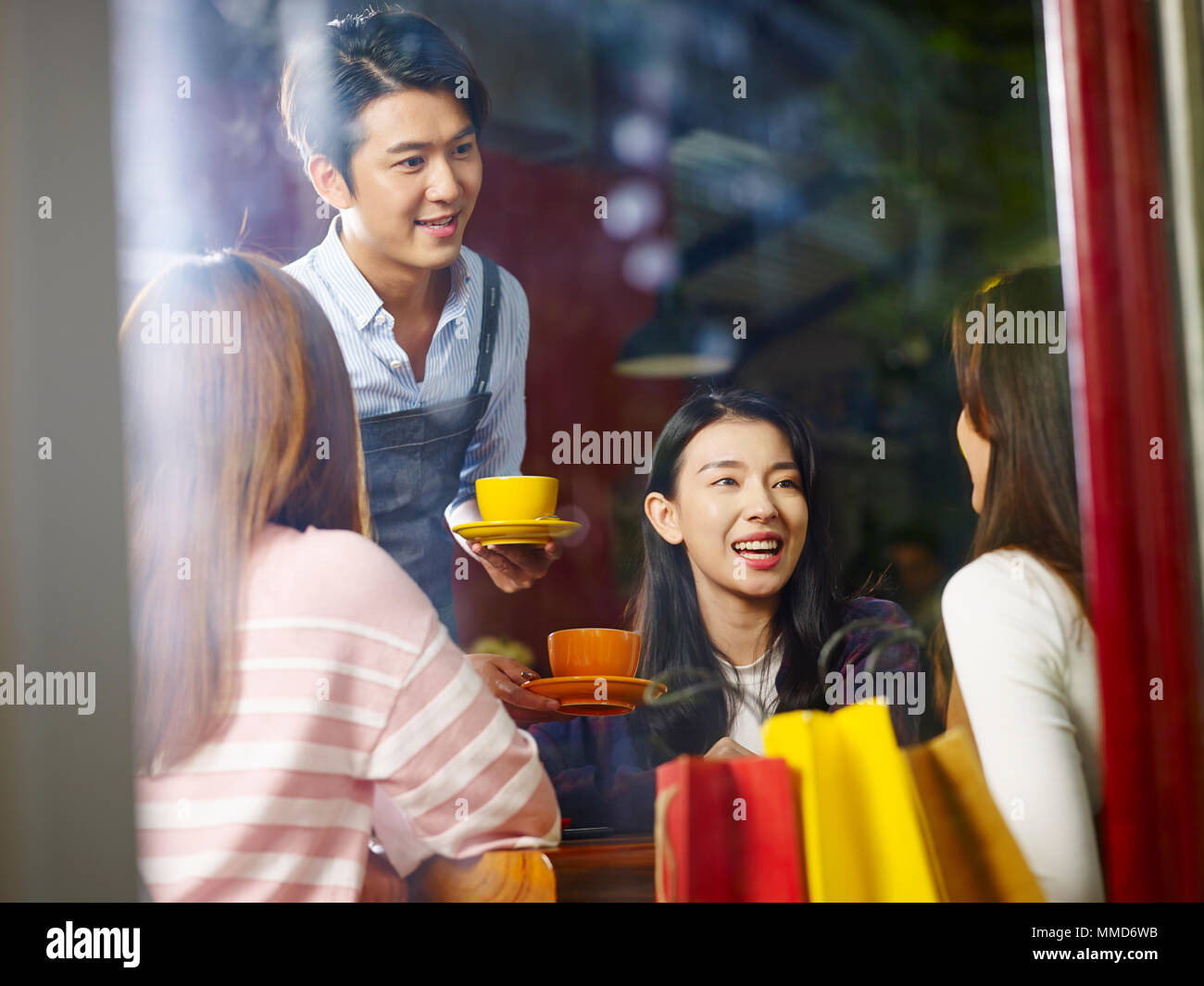 Giovani asiatici cameriere che serve i clienti di sesso femminile nel coffee shop, shot attraverso il vetro di una finestra. Foto Stock