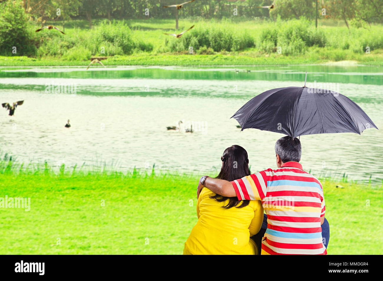 Vista posteriore coppia Senior seduta di erba sotto ombrellone vicino al lago At-Park Foto Stock