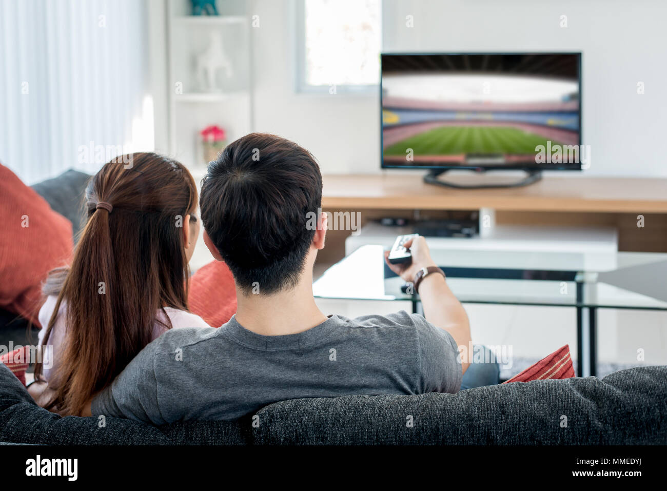 Vista posteriore della coppia asiatica guardando la partita di calcio in televisione nel soggiorno. Festa del calcio concetto. Foto Stock