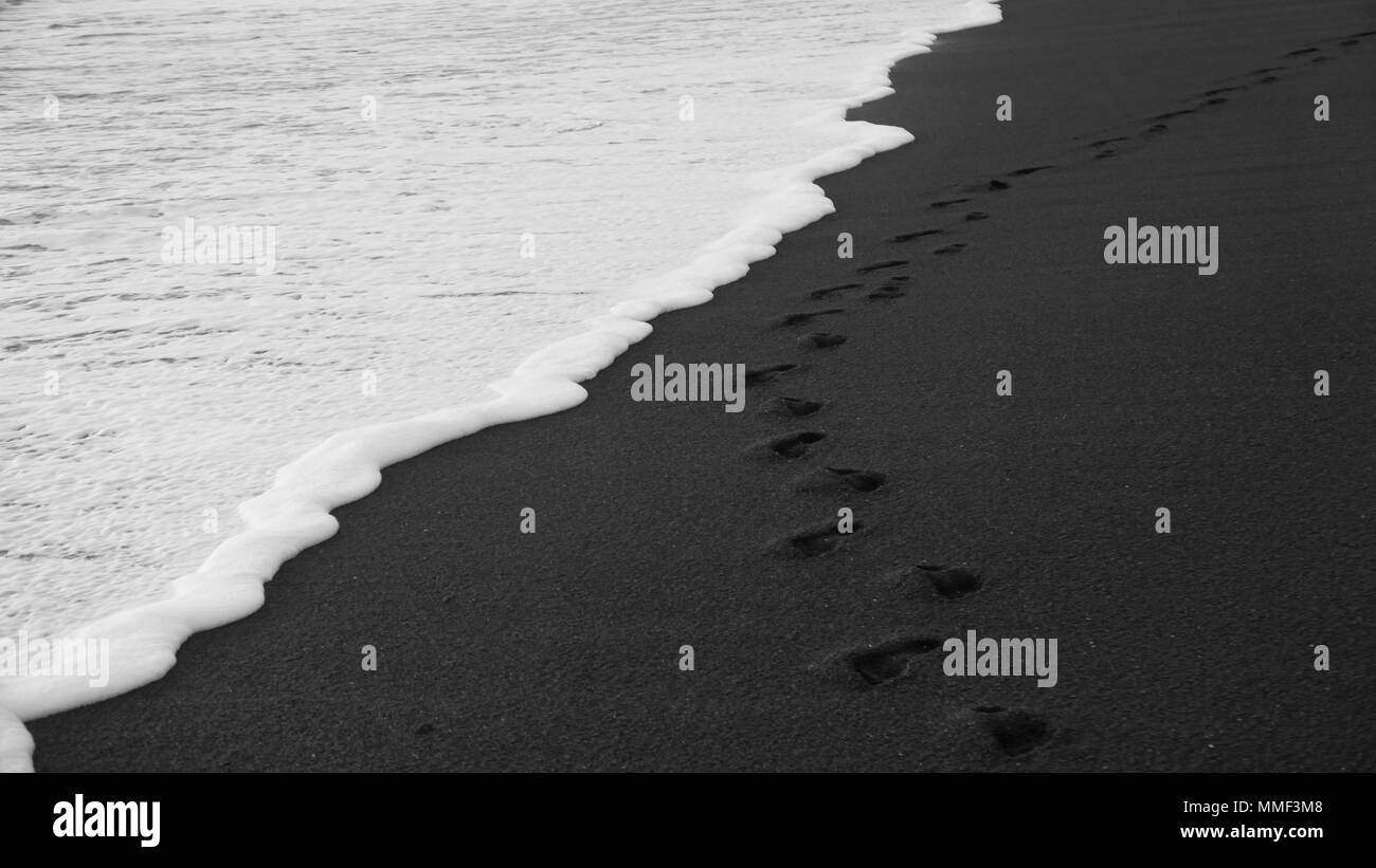 Mare Bianco di schiuma acqua toccando impronte sulla spiaggia di sabbia nera Foto Stock