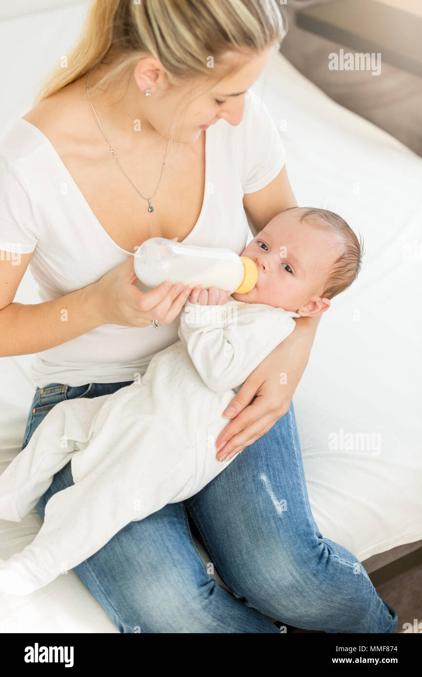 Bella giovane donna seduta sul letto e alimentando il suo bambino da bottiglia Foto Stock