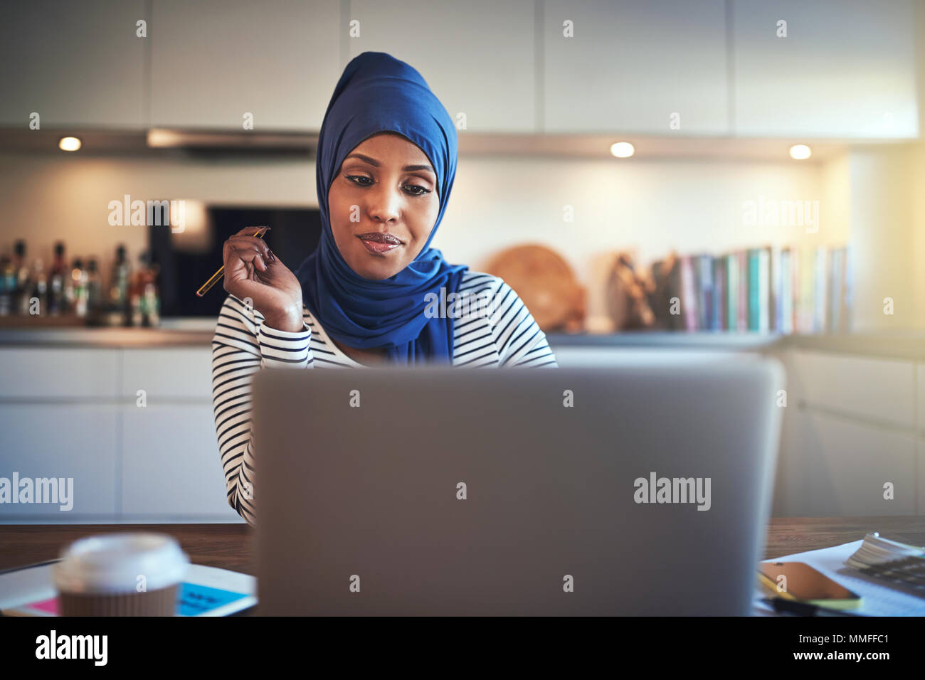 Sorridente giovane musulmano imprenditore femmina indossando un hijab seduti a un tavolo nella sua cucina a scrivere delle note e al lavoro su un notebook Foto Stock