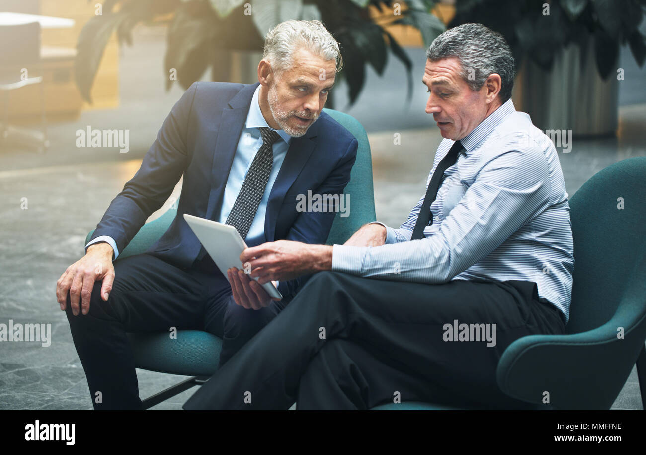 Due ben vestito imprenditori matura seduti nelle sedie insieme in un ufficio lobby e parlare insieme e utilizzando una tavoletta digitale Foto Stock