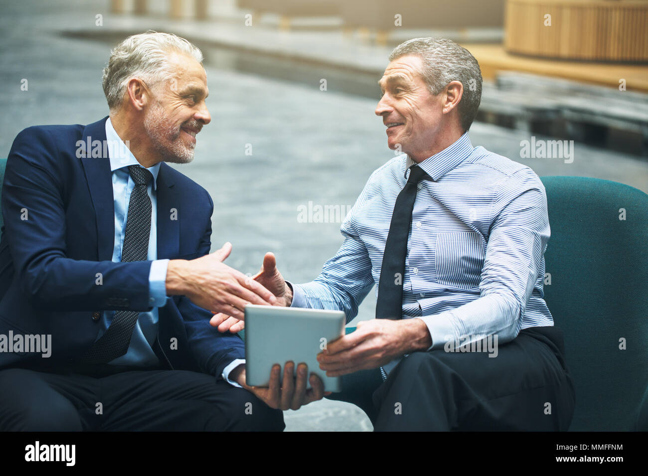 Due sorridenti imprenditori matura seduti su sedie in un ufficio lobby e agitando le mani insieme e utilizzando una tavoletta digitale Foto Stock