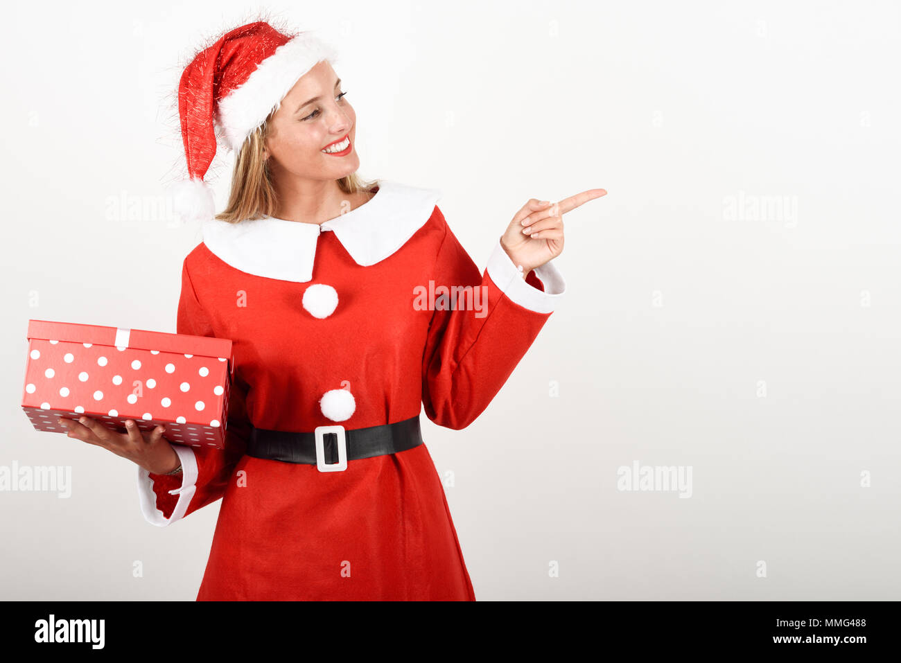 Donna bionda in Santa Claus vestiti sorridente con confezione regalo nelle sue mani. Giovani femmine con gli occhi blu rivolta a destra con le sue dita, isolato su bianco Foto Stock