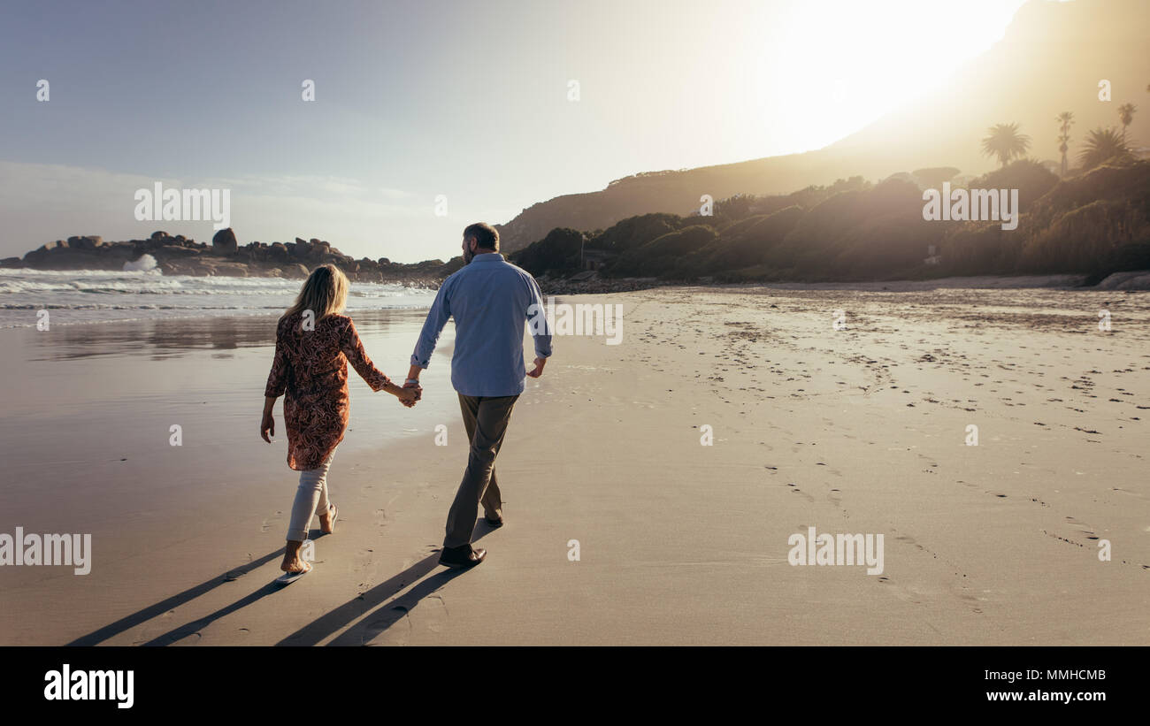 Vista posteriore della romantica coppia senior camminando mano nella mano sulla spiaggia. Uomo maturo e la donna facendo una passeggiata lungo la riva del mare. Foto Stock