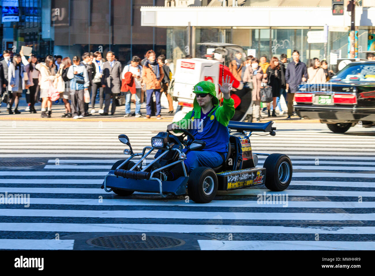 Tokyo, Shibuya crossing. Popolari attività turistiche, guida MariCar, Mario Kart mentre vestita come Mario carattere. L'uomo sventolando durante la guida. Foto Stock