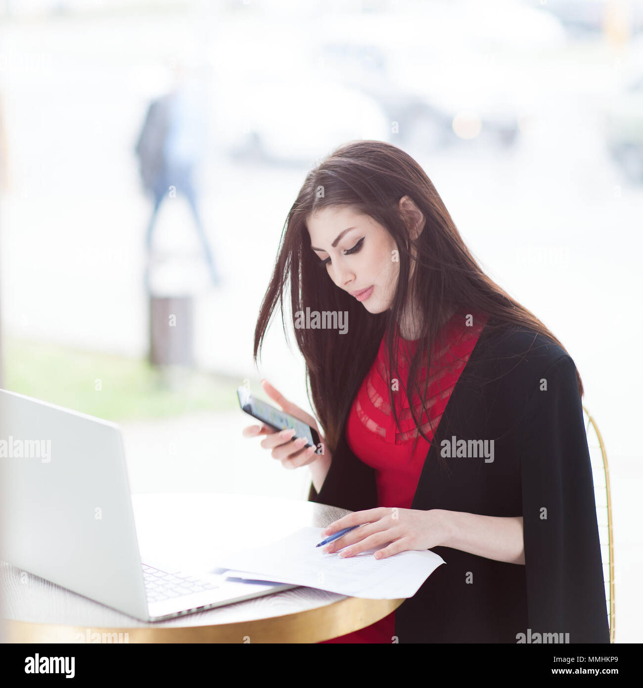 Bel giovane donna con il cellulare in mano in seduta cafe con ampia finestra e studiano documenti udienza dal computer portatile Foto Stock