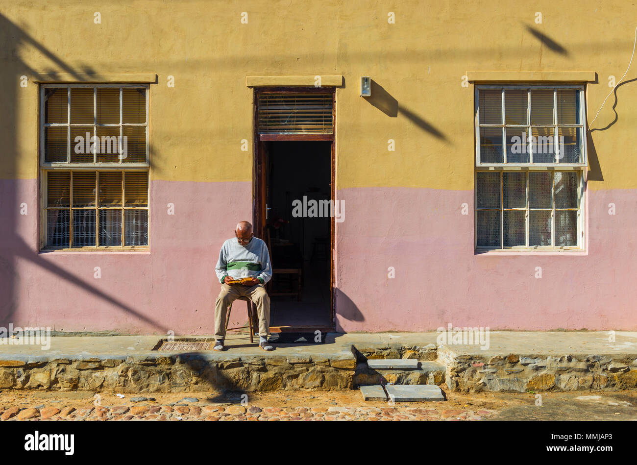 Un anziano uomo adulto seduto su un marciapiede e la lettura di un libro di fronte a una facciata colorata nel quartiere Malese di Bo Kaap a Cape Town, Sud Africa. Foto Stock