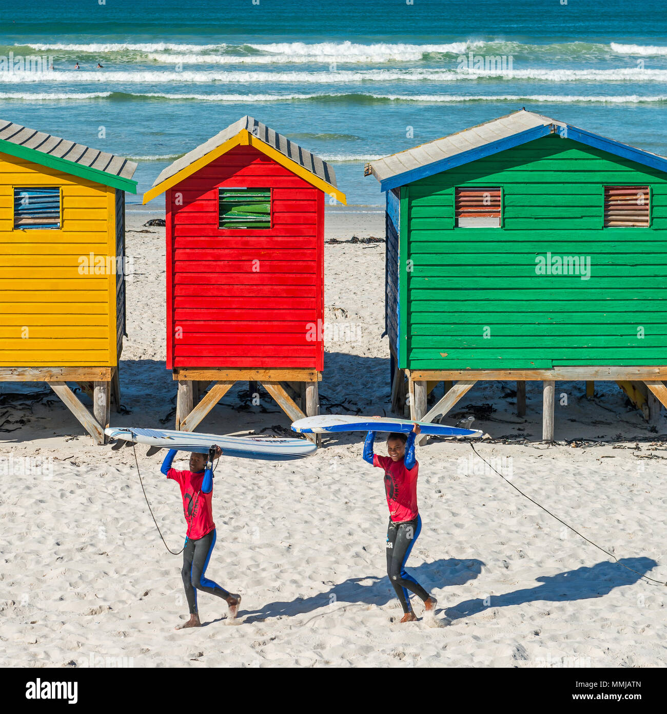Sorridente giovane sudafricano surfers che trasportano le loro tavole da surf sulla spiaggia di Muizenberg, famoso per la pittoresca spiaggia di capanne, Sud Africa. Foto Stock