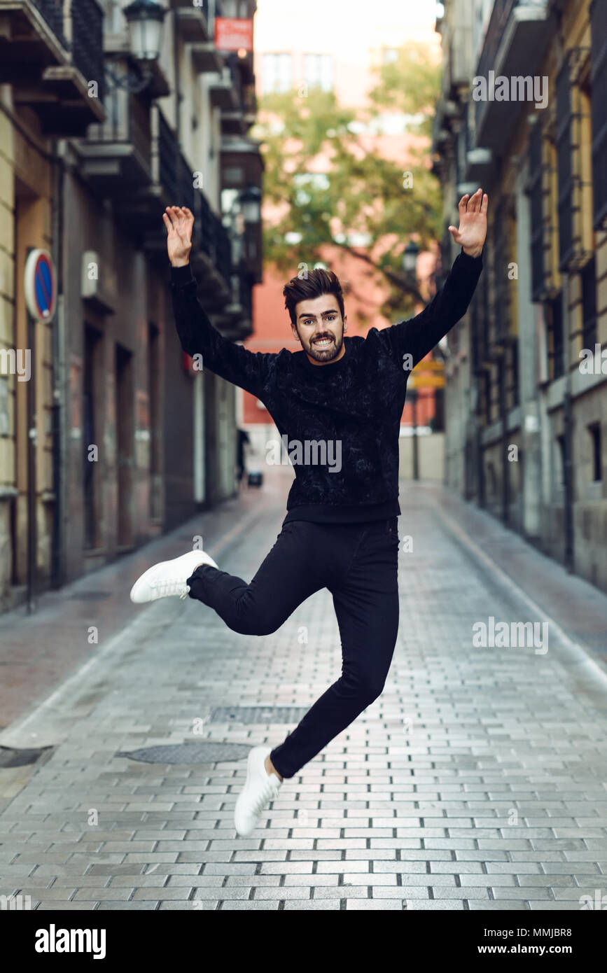 Giovane uomo barbuto jumping in background urbano con le braccia aperte di indossare un abbigliamento informale. Ragazzo con la barba e acconciatura moderna in strada. Foto Stock