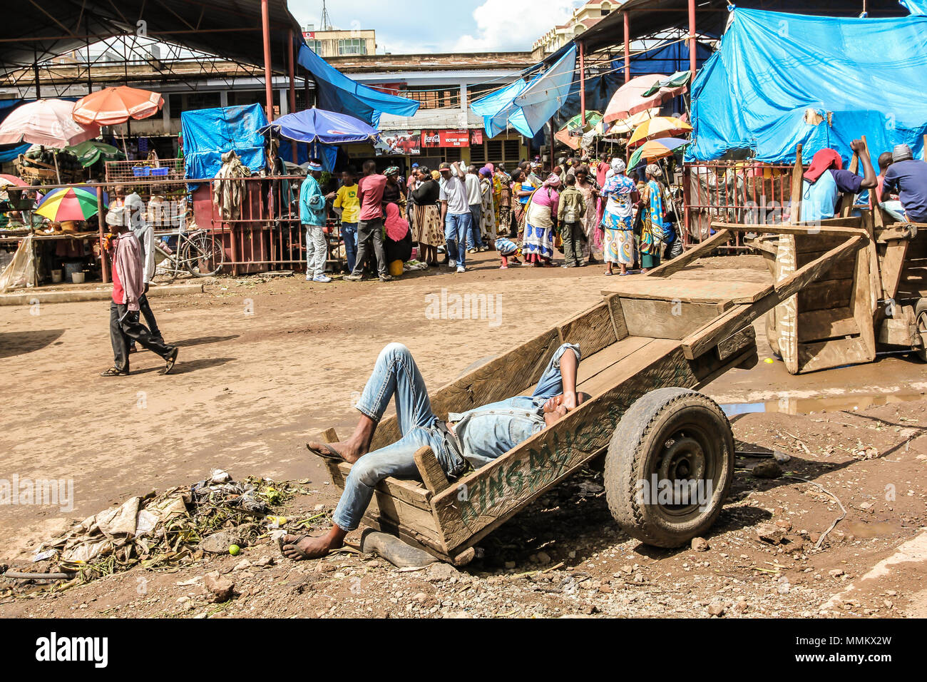 Arusha, Tanzania Africa - Gennaio 2, 2013: uomo disteso su un carrello di legno dorme in un mercato della città. Questi mercati africani sono pieni di colori e di persone che vendono i loro prodotti locali e loro coltivano frutta e verdura Foto Stock