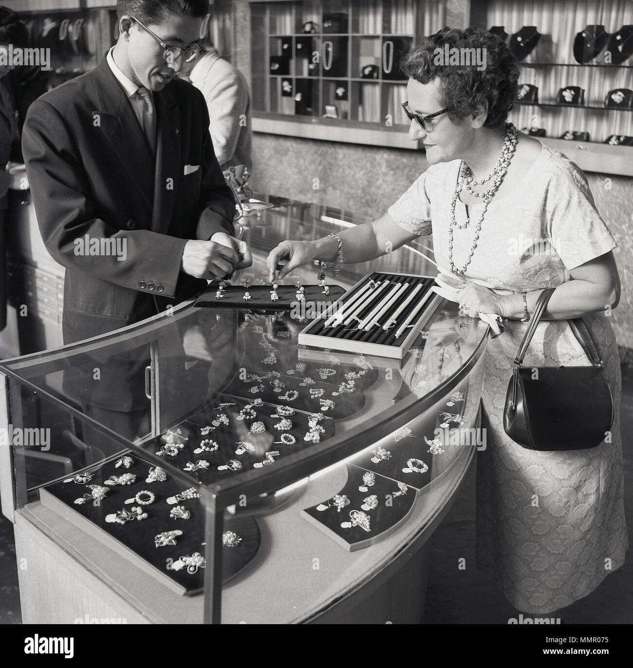 Degli anni Cinquanta, storico western signora con borsetta in  corrispondenza di un banco espositore all'interno di un negozio gioielli  essendo mostrati alcuni gioielli perla da un giapponese assistente vendite,  Tokyo, Giappone. Il