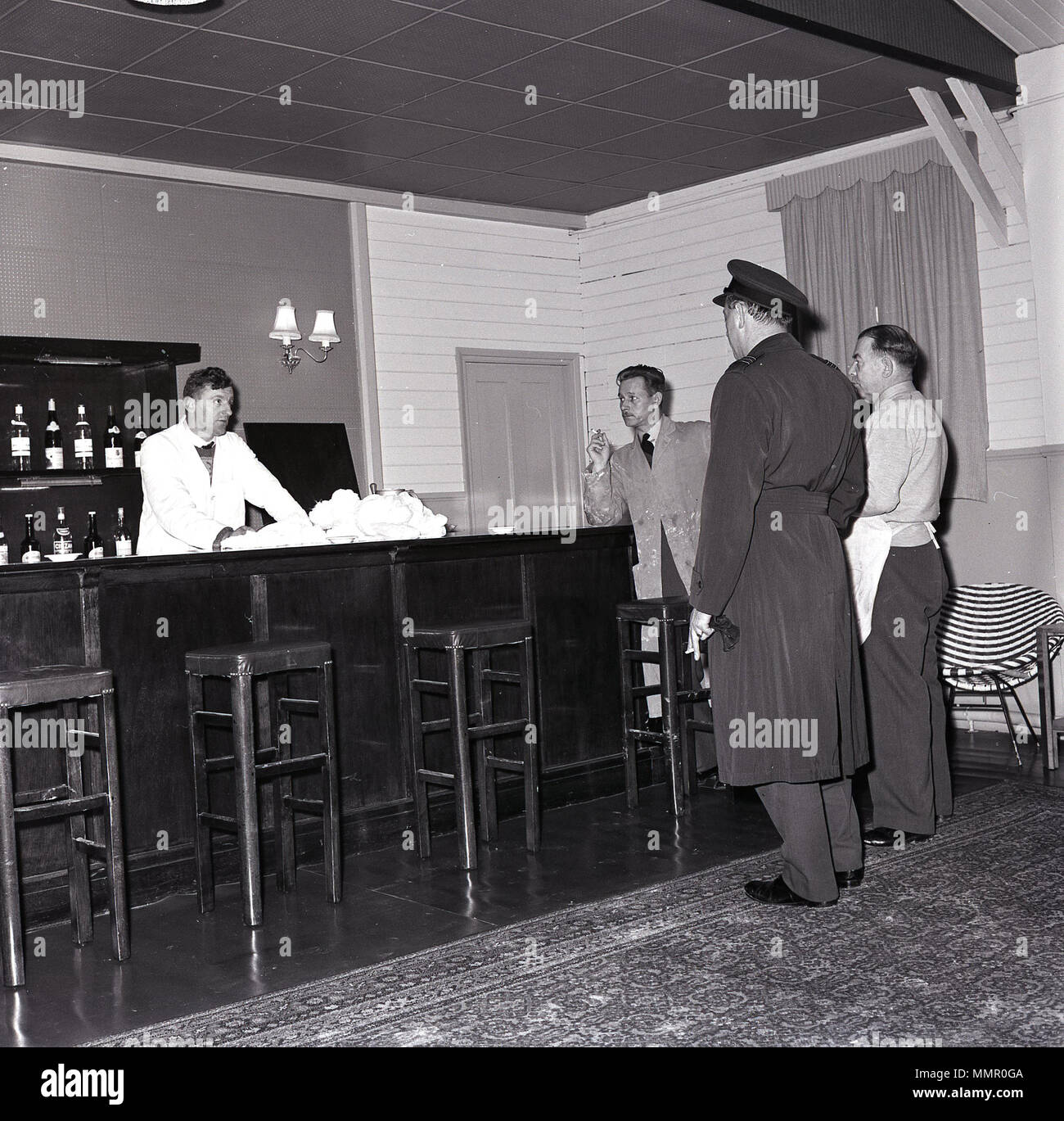 1965, RAF officer in cappotto e cappello con personale di catering in piedi al bar teatro a RAF Halton, Aylesbury, Inghilterra, Regno Unito avente una sigaretta. Foto Stock