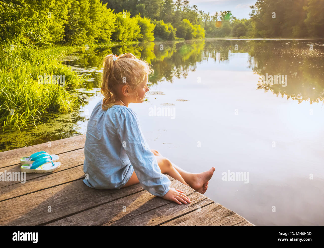 Lonely piccola ragazza sogna sul dock vicino al piccolo lago sul caldo giorno d'estate Foto Stock