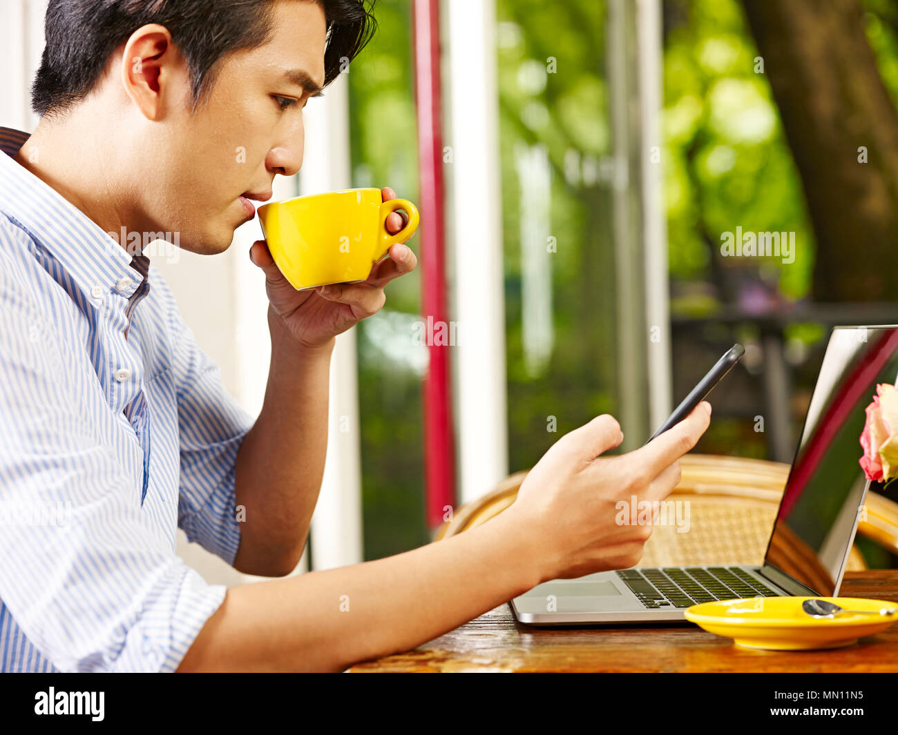 Giovane uomo asiatico mediante telefono cellulare e computer portatile mentre si beve il caffè cappuccino all'aperto Foto Stock