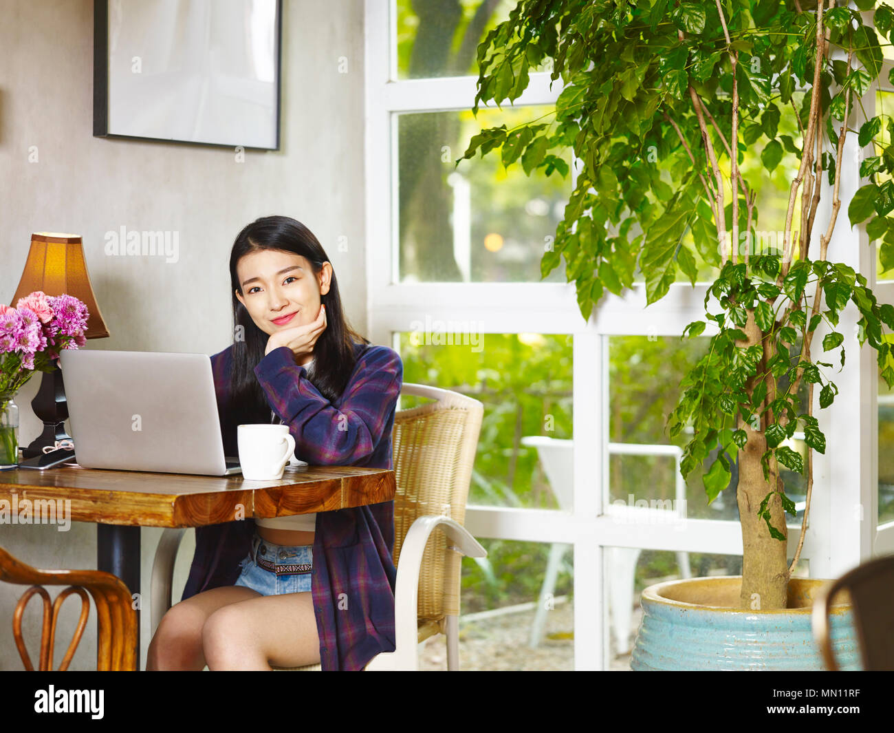 Bella giovane donna asiatica guardando fotocamera a sorridere mentre si utilizza il computer portatile in una caffetteria. Foto Stock