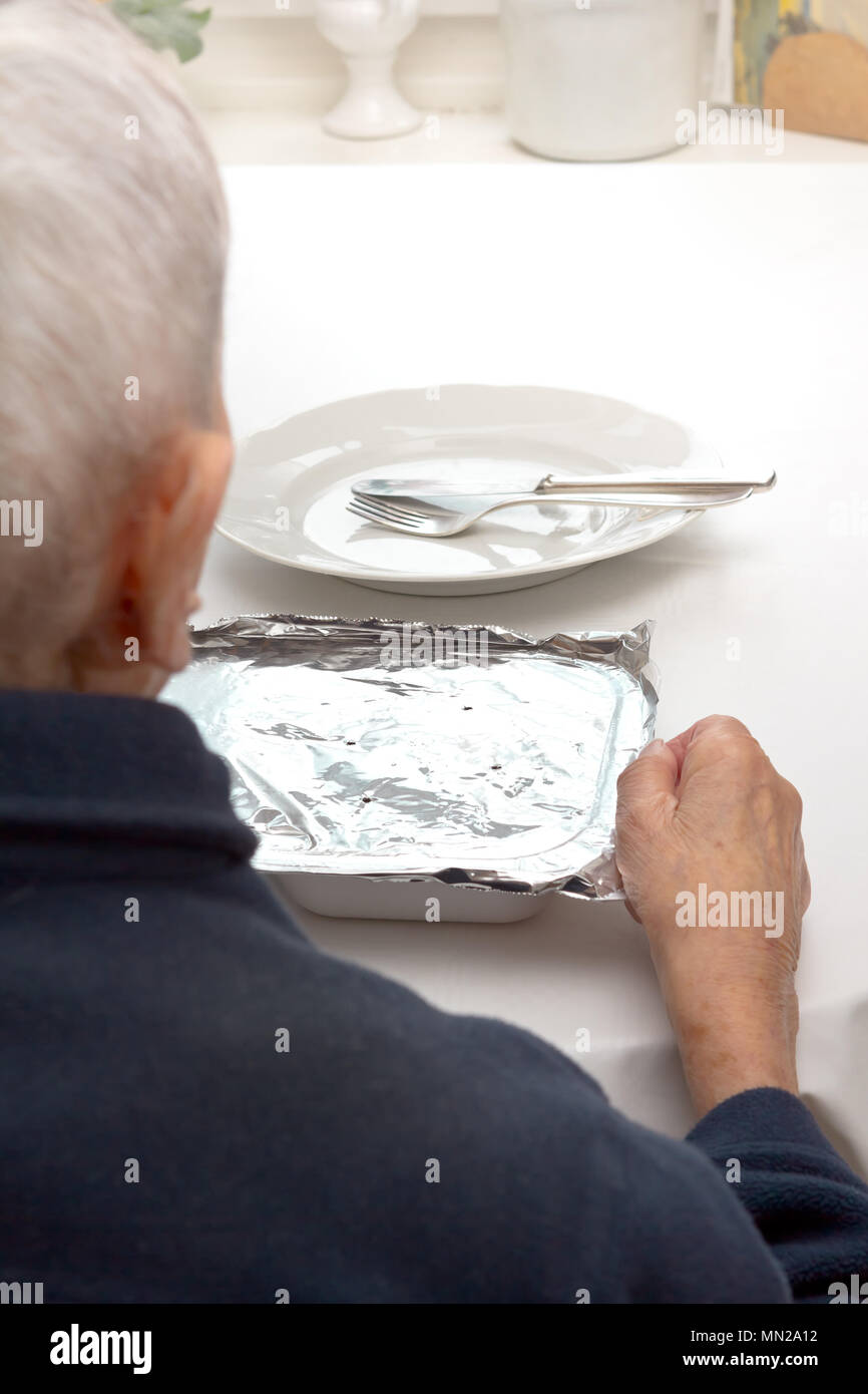 Il vecchio donna seduta da sola a un tavolo nella sua cucina o soggiorno con piastra, posate e un pasto precotto da alimentare un servizio di consegna Foto Stock