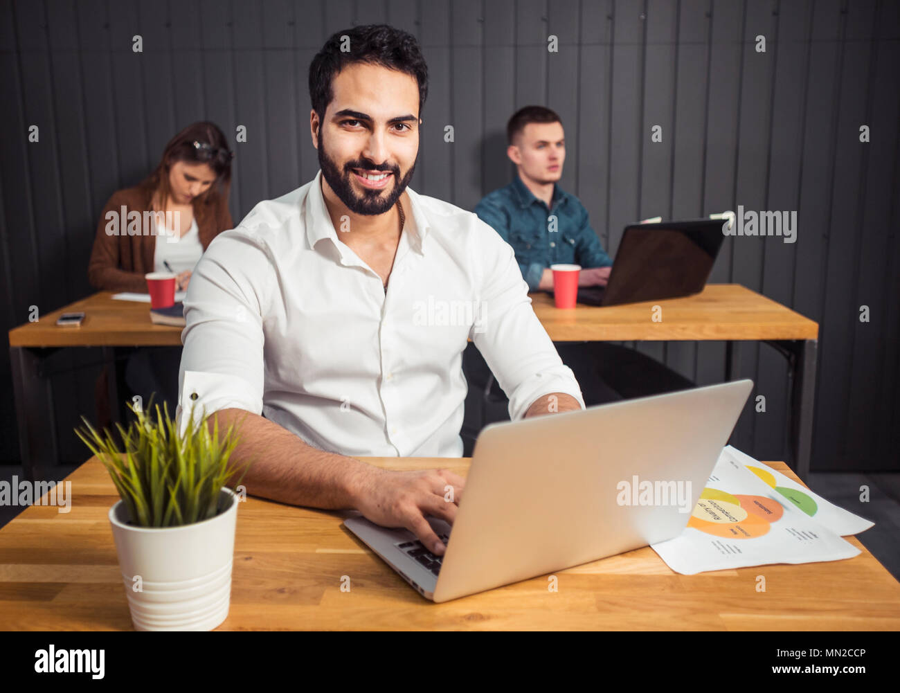 Uomo freelance con barba nera lavorando sul computer portatile prima di colleghi a lavorare sull'attività, preparando la prossima fase del progetto alla scrivania da ufficio con grafici Foto Stock