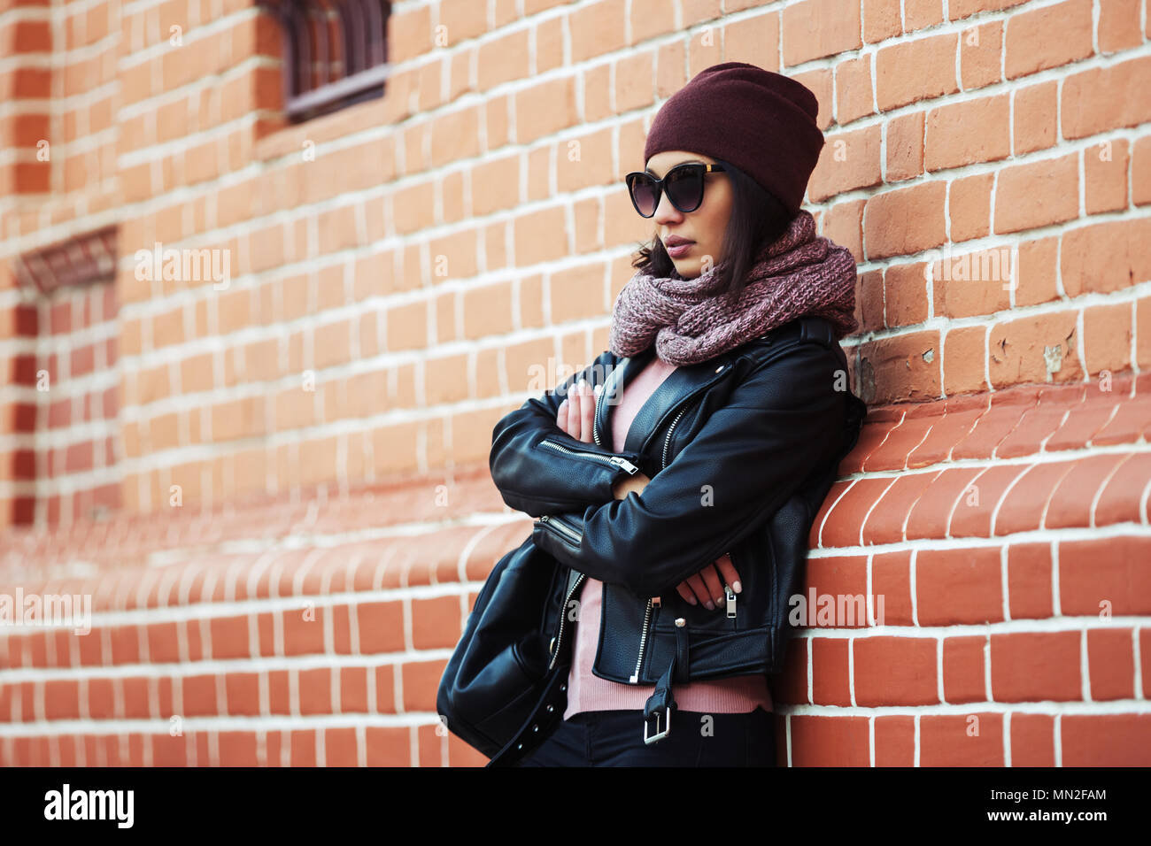 Moda giovane donna che indossa gli occhiali da sole e sciarpa beanie appoggiato su di un muro di mattoni elegante modello femminile in pelle nera camicia outdoor Foto Stock