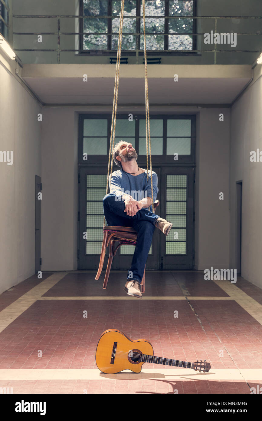 Uomo seduto su una sedia pensile e una chitarra sul pavimento, interno Foto Stock