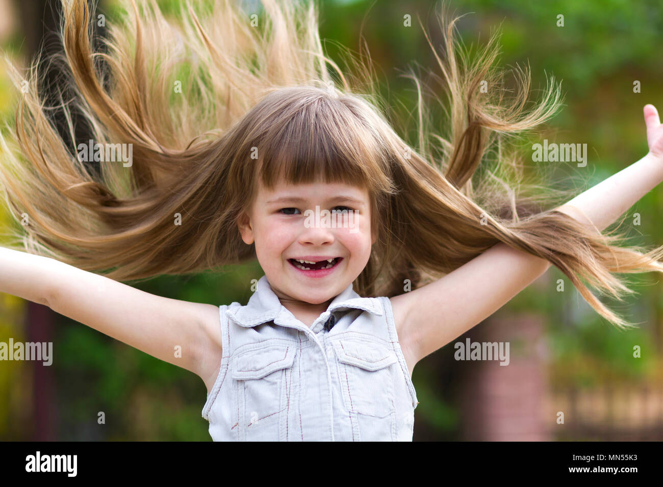Graziosa ragazza in età prescolare in vestito bianco senza maniche e con  bellissimi capelli lunghi biondi soffiata dal vento, divertente sorriso  priva di denti e diffondere bracci su sfocato Foto stock -