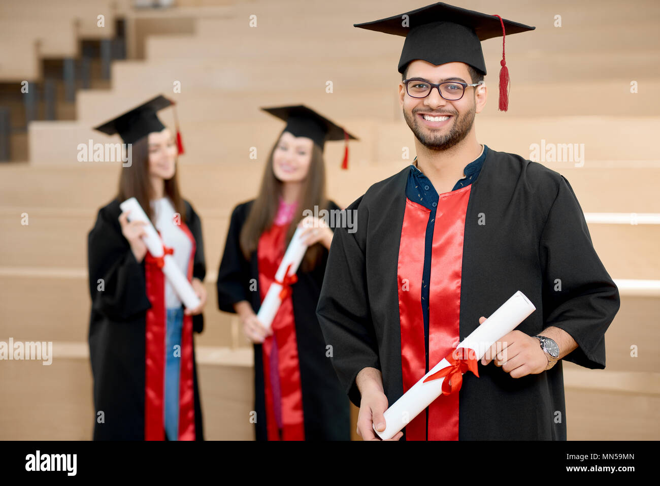 Sorridenti boy mantenendo il diploma universitario. Laureato in piedi vicino a giovani groupmates e guardando soddisfatto. Gli studenti vestita di nero e colorato di rosso abiti di graduazione. Terminando l'università. Foto Stock