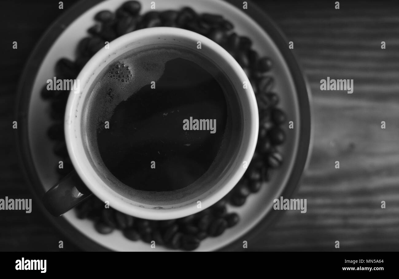 Una tazza di caffè caldo su un piattino con un chicco di caffè. Sullo sfondo di una tavola di legno. Texture, sfondo. Foto in bianco e nero Foto Stock