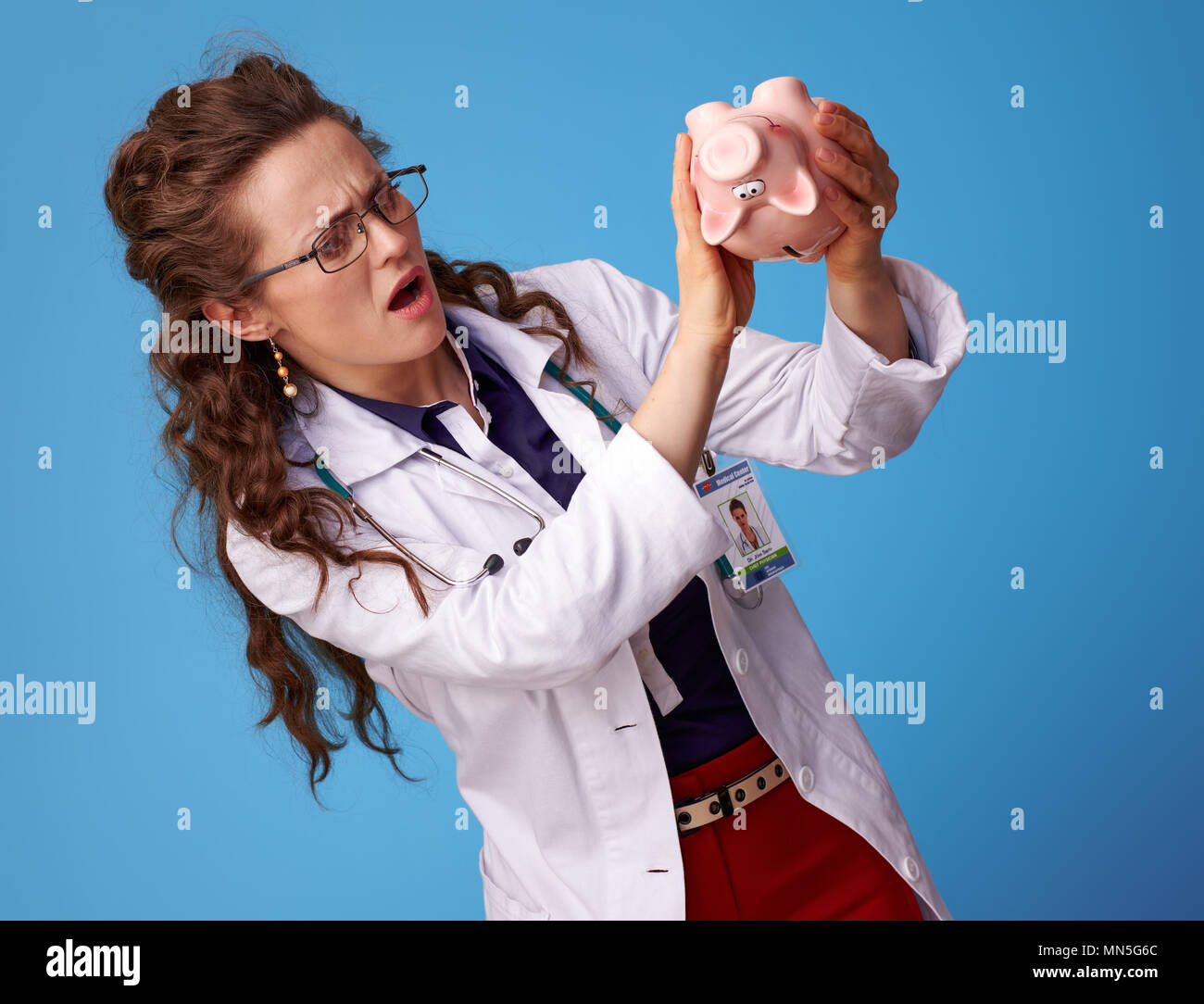 Sorridente medico donna in bianco manto medico lo scuotimento di un maiale salvadanaio su sfondo blu Foto Stock