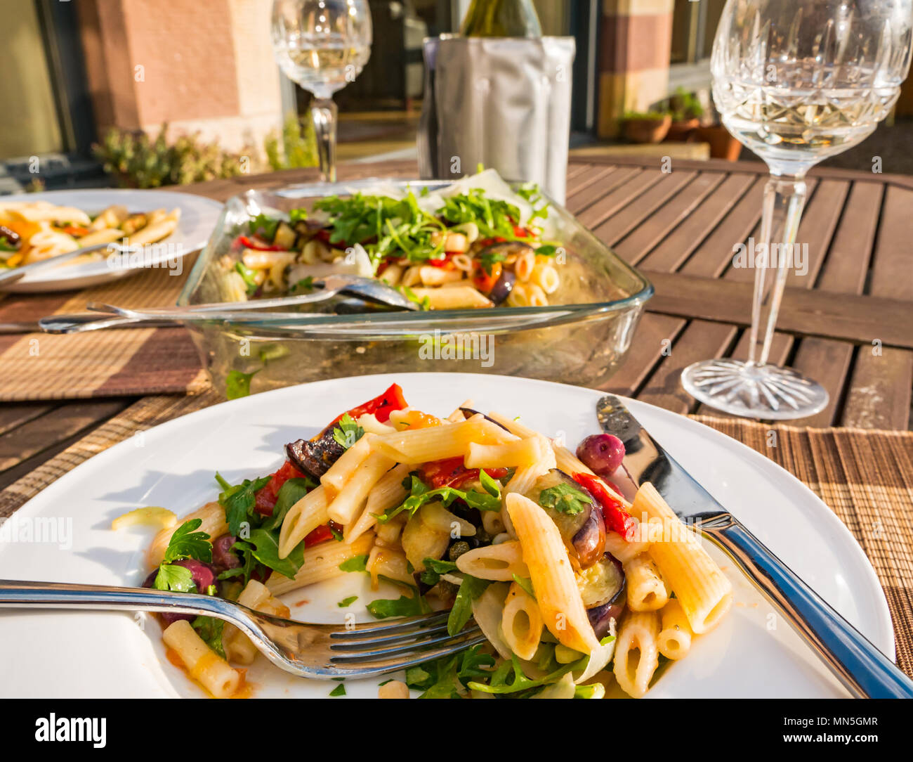 Tavolo esterno nella luce del sole con insalata di pasta piatto servito su bianco cena piastra, crystal bicchieri di vino e una bottiglia di vino bianco freddo nello scambiatore di calore Foto Stock