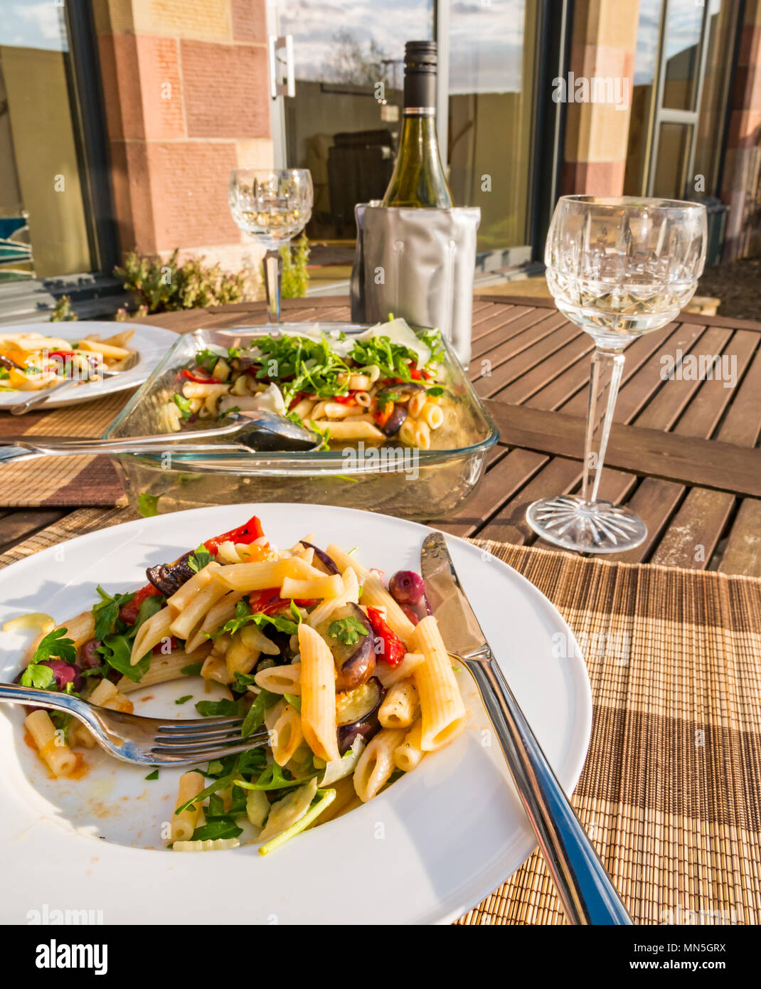 Tavolo esterno nella luce del sole con insalata di pasta piatto servito su bianco cena piastra, crystal bicchieri di vino e una bottiglia di vino bianco freddo nello scambiatore di calore Foto Stock