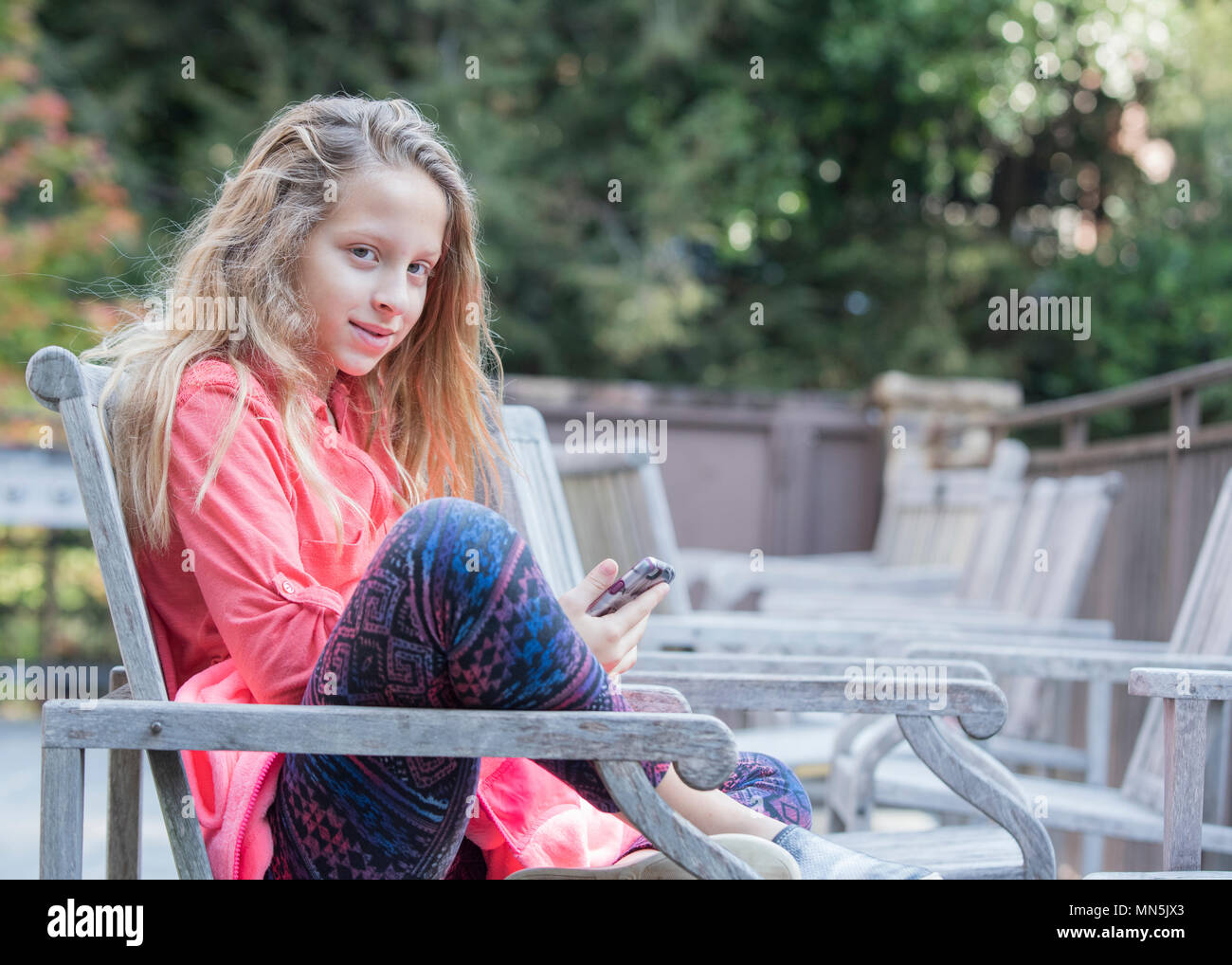 Ragazza giovane seduto in una sedia al di fuori con il suo telefono cellulare. Ella è rilassato e guardando leggermente di lato alla fotocamera. Foto Stock