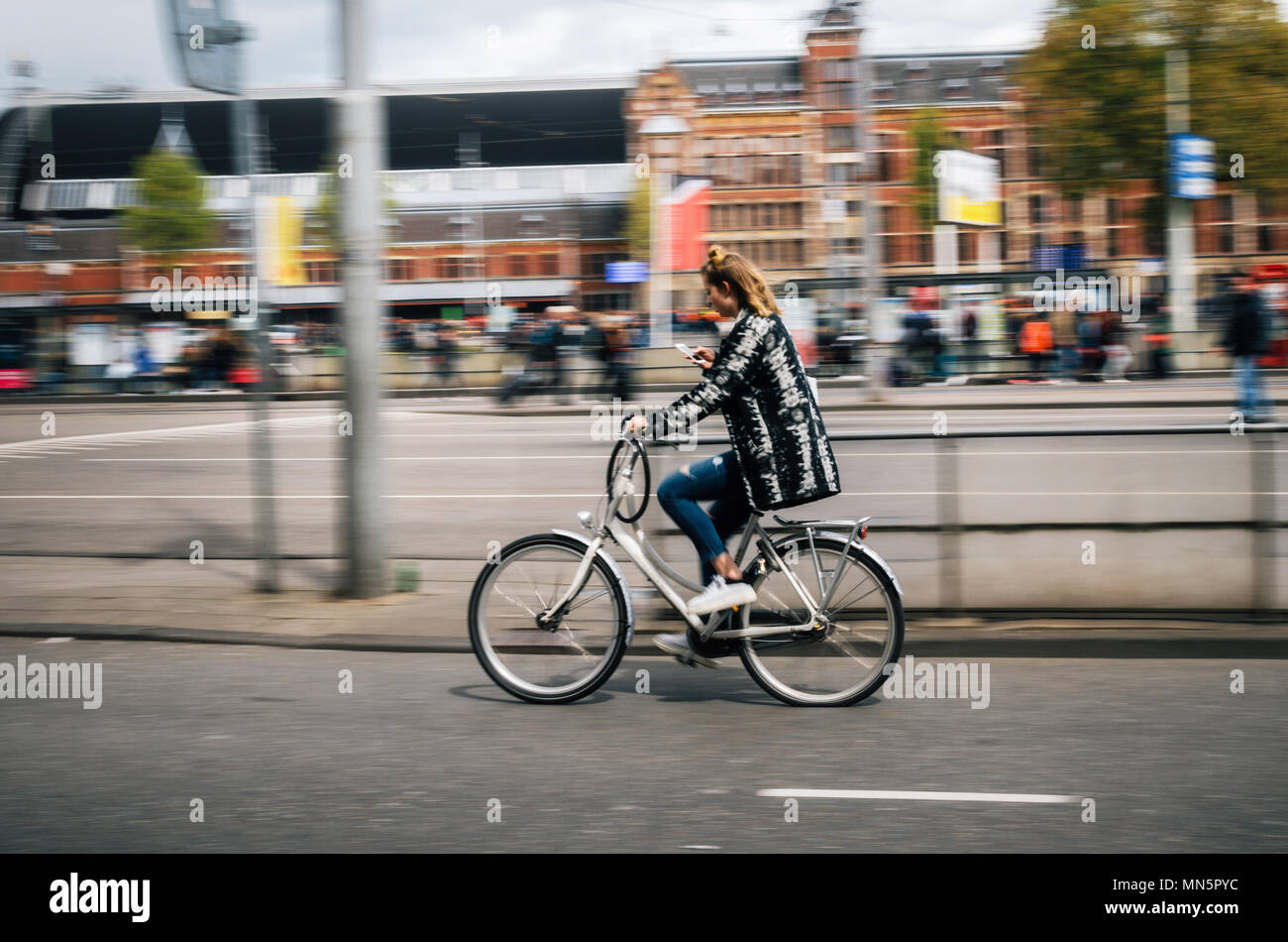 Amsterdam, Paesi Bassi - 27 April, 2017: giovane donna corse in bicicletta e guarda nel telefono cellulare tenendo in una mano. Strade di Amsterdam, Paesi Bassi. Mo Foto Stock