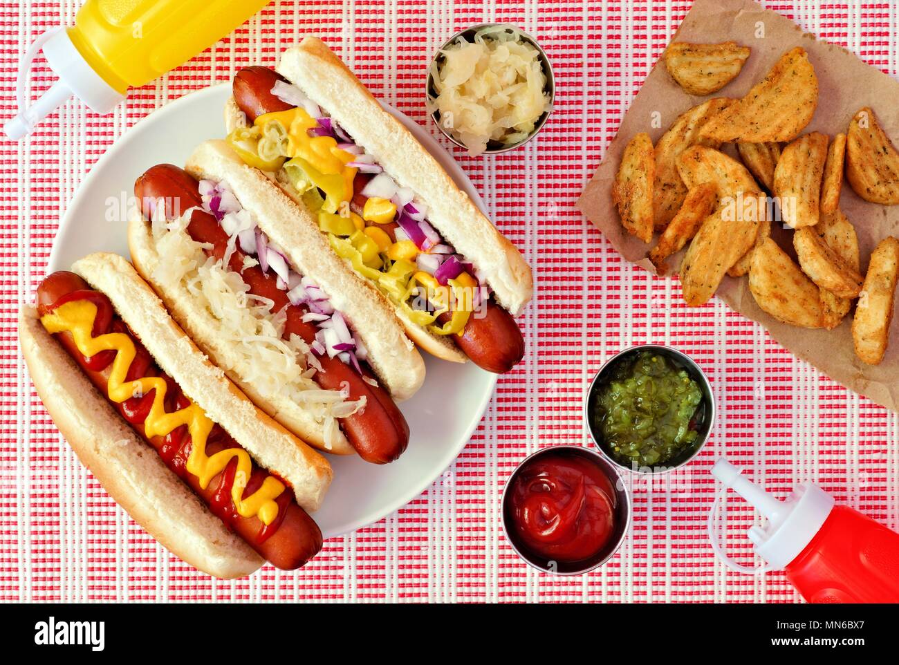 Hot Dogs con assortimento di condimenti e Spicchi di patate al di sopra di scena su un rosso e un panno bianco Foto Stock