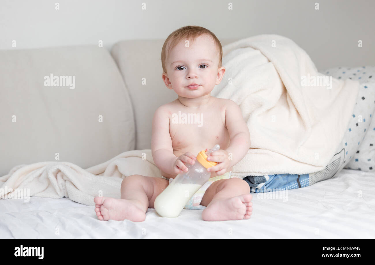 Adorable baby boy in pannolini seduta sul letto e tenendo la bottiglia di latte Foto Stock