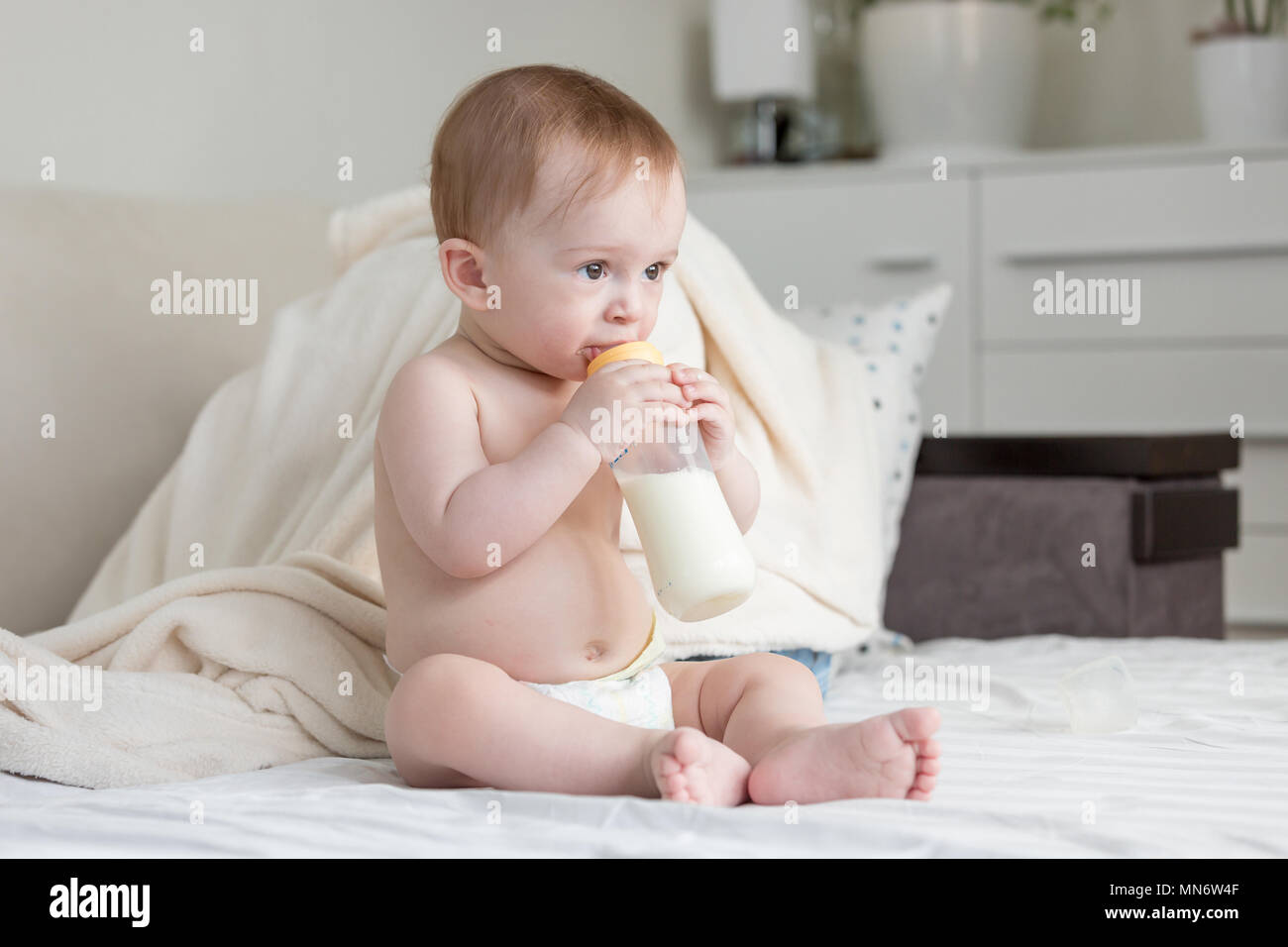 Ritratto di simpatici baby boy in pannolini seduta sul letto e bere latte dalla bottiglia Foto Stock