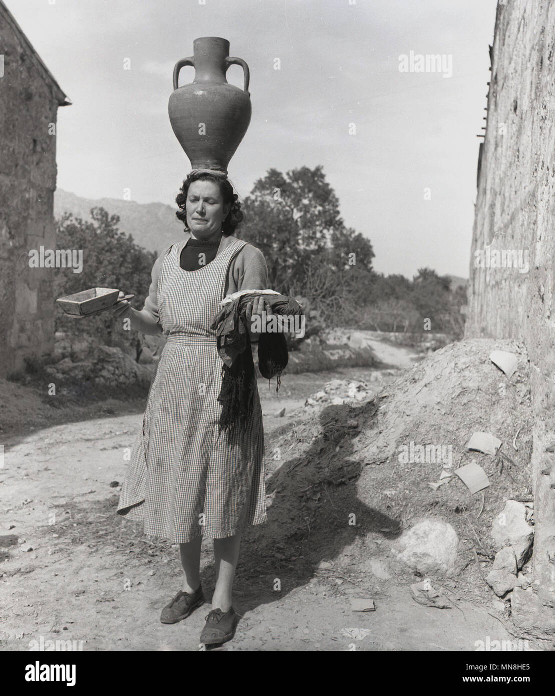 Degli anni Cinquanta, foto storiche, un villaggio di Maiorca, donna in piedi su un percorso rurale visualizzazione eccezionale capacità di bilanciamento, con una grande vetrata pentola di creta sul suo capo e prodotti locali in ogni mano, Maiorca, SPAGNA, Foto Stock