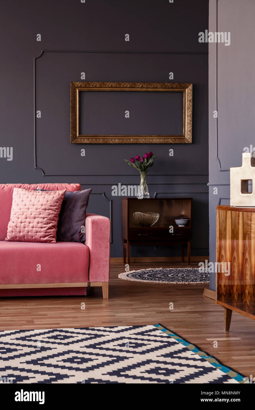 Cuscini decorativi sul lettino rosa in piedi nel buio salotto interno con fiori freschi, moquette a motivi geometrici e armadio vintage Foto Stock