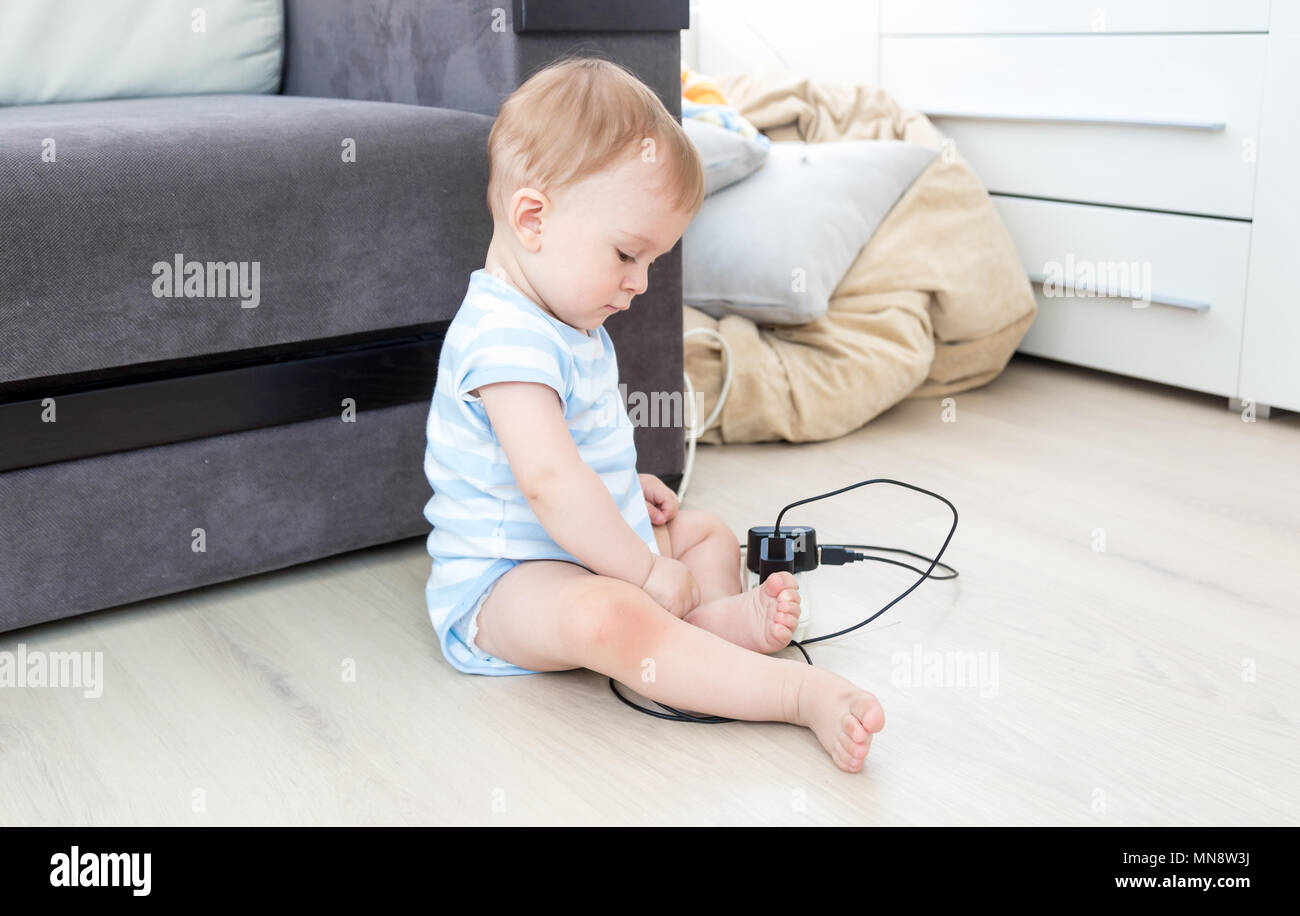 9 mesi di età bambino seduto sul pavimento nel soggiorno e giocando con le prese di corrente e cavi elettrici Foto Stock