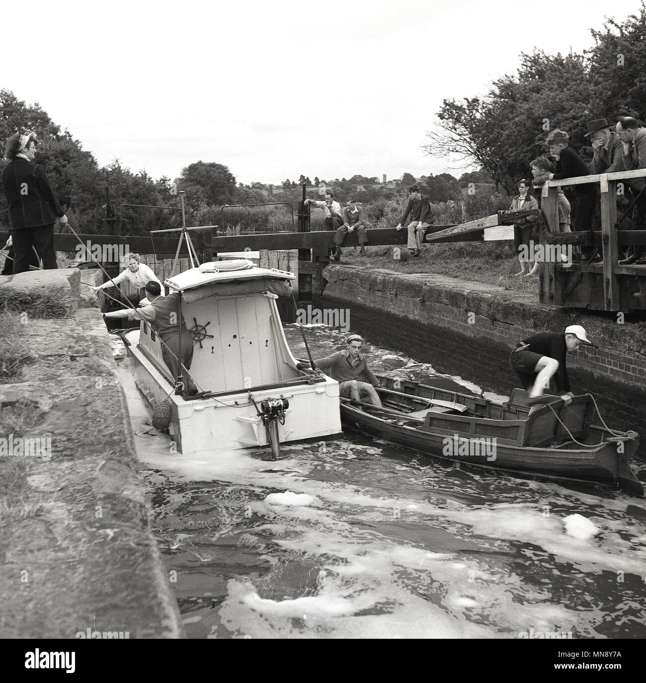 Degli anni Cinquanta, storico, persone a guardare come un piccolo motoscafo e un di legno barca a remi tentare di manovra che passano ogni altro su un canale rurale bloccare, Inghilterra, Regno Unito. Foto Stock