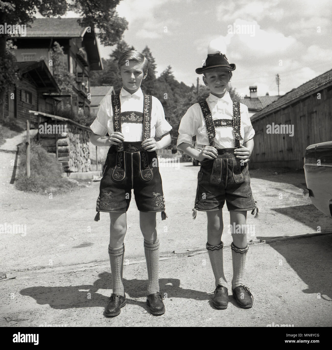 Degli anni Cinquanta, i due giovani ragazzi bavarese di stare in piedi insieme su una pista polverosa al di fuori di un villaggio delle Alpi, Germania, indossando abiti tradizionali del SER, breve pantaloni di pelle o lederhosen, Foto Stock