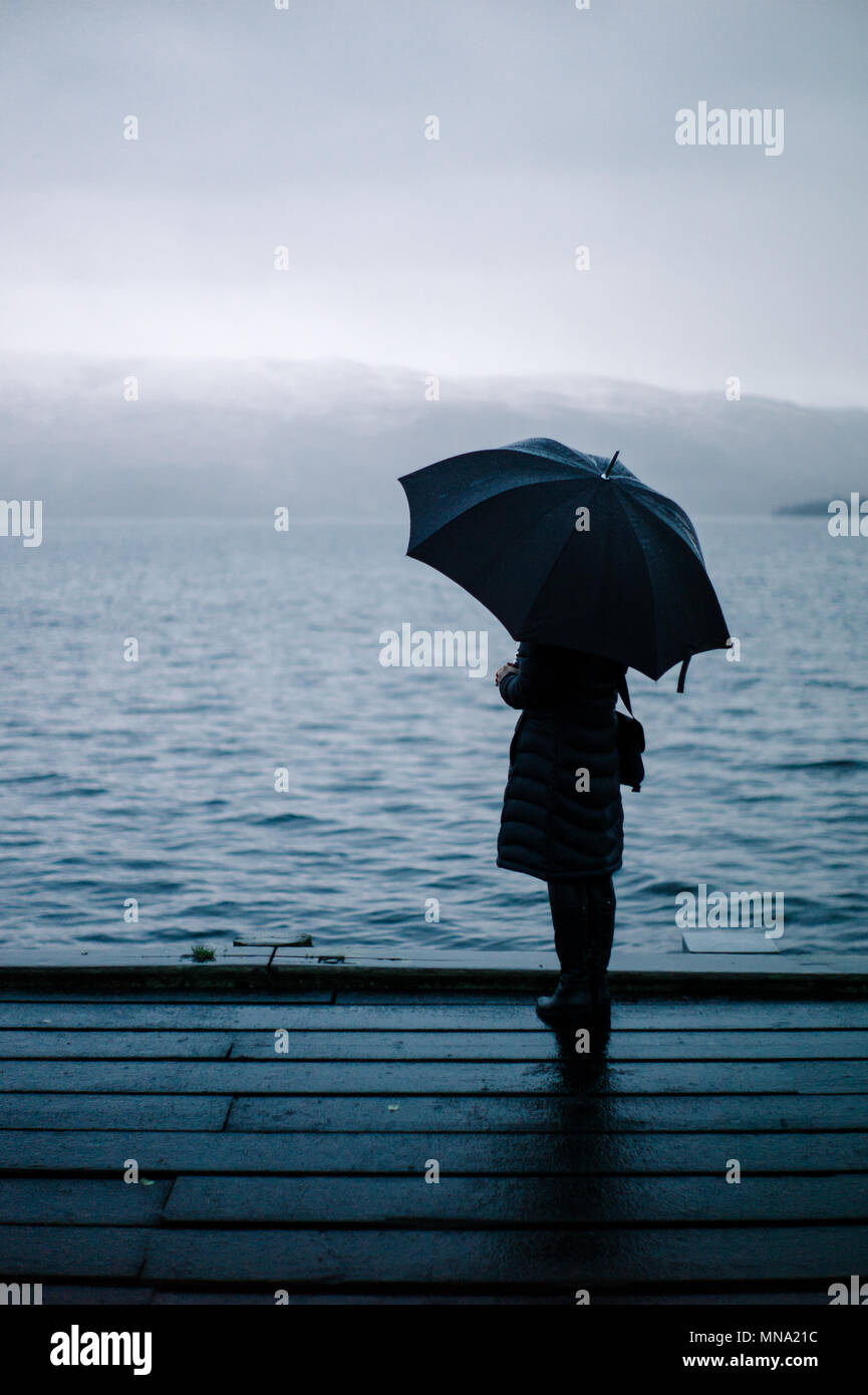 Donna che guarda fuori sopra Loch Lomond con un ombrello in una nebbiosa inverni freddi giorno. Foto Stock