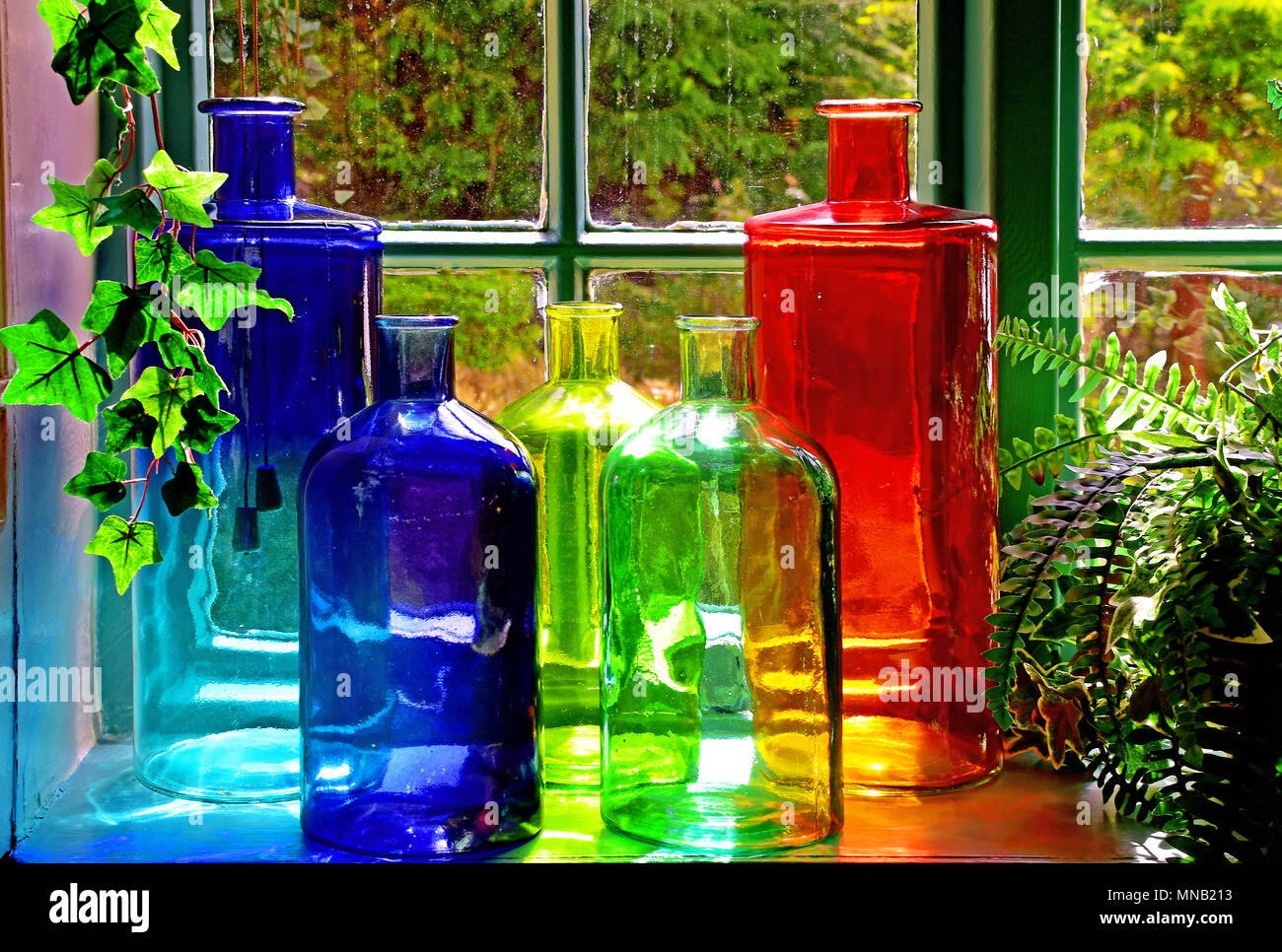 Rosso verde giallo e azzurro bottiglie dalla finestra con felci ed edera Foto Stock