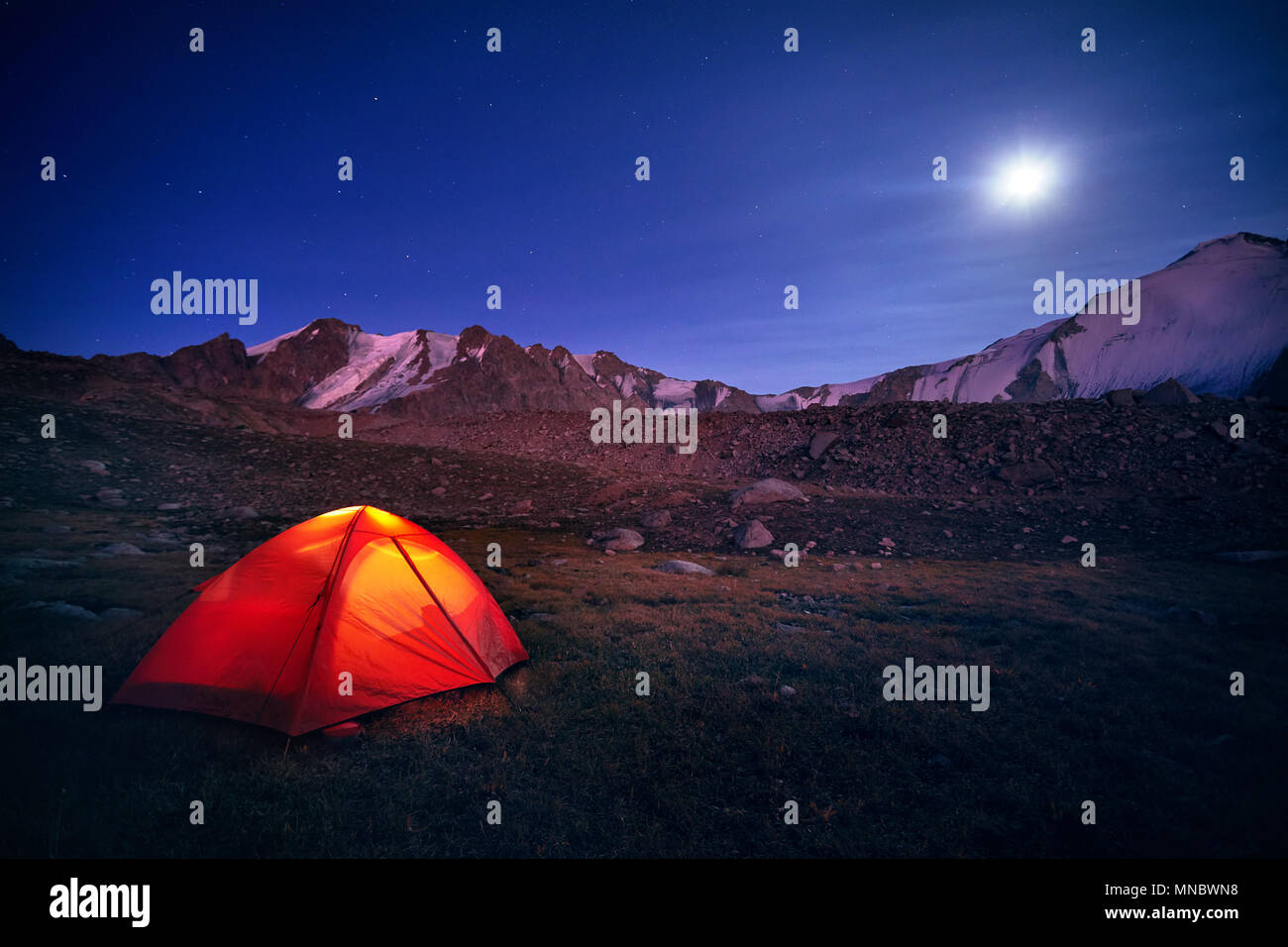 Tenda arancione si accende sotto il cielo notturno pieno di stelle e la luna in montagna in Kazakistan Foto Stock