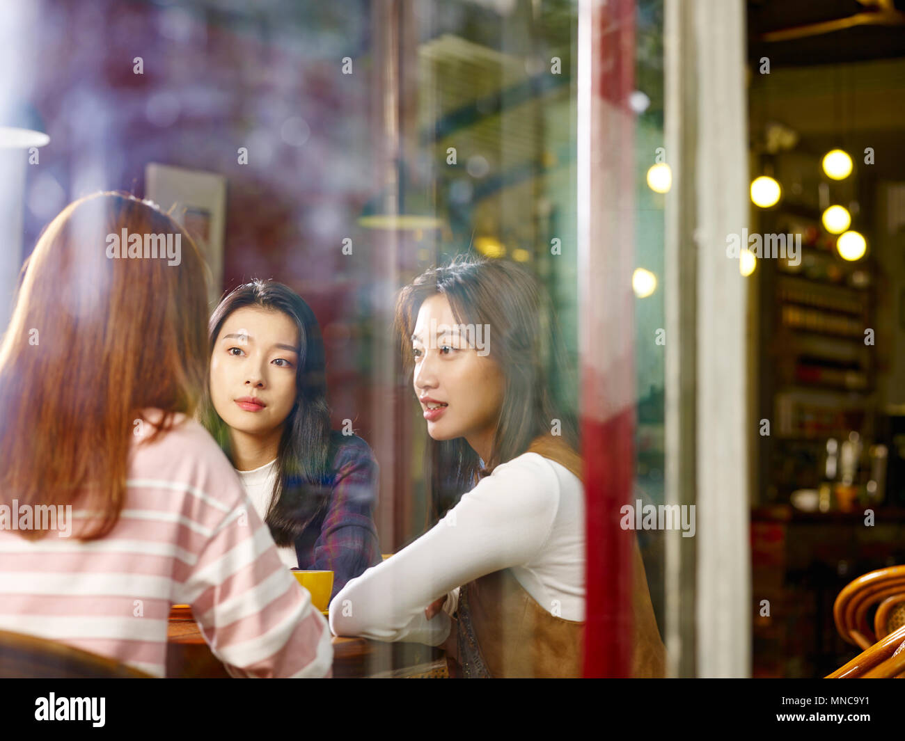 Tre felice giovane e belle donne asiatiche seduta a tavola in chat parlando in negozio di caffè o tè casa, shot attraverso il vetro di una finestra. Foto Stock