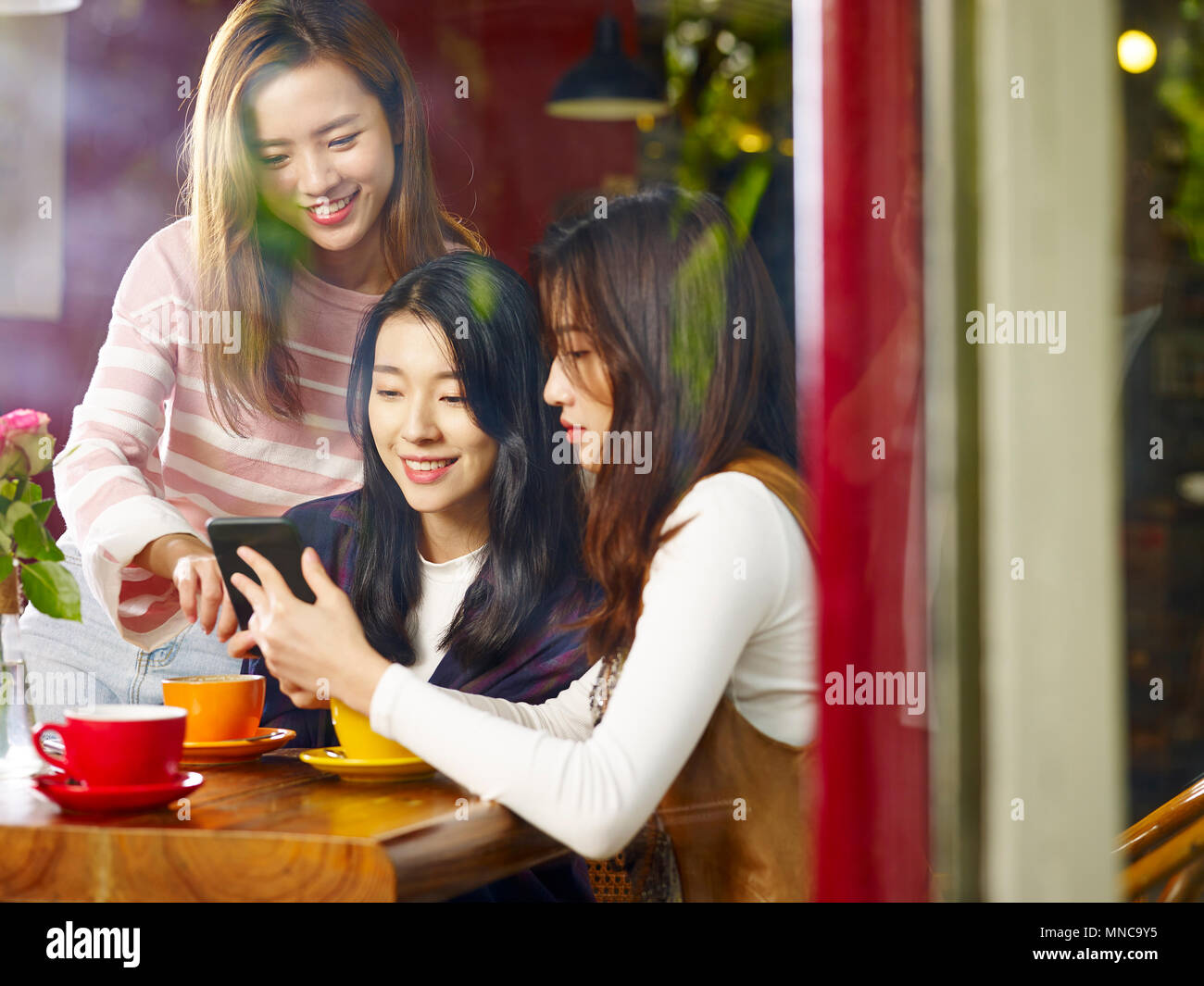 Tre felice giovane e belle donne asiatiche seduta a tavola in chat parlando a giocare con il cellulare nel negozio di caffè o tè casa, shot attraverso la finestra glas Foto Stock