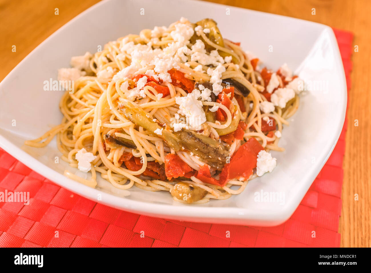 Un piatto bianco su un posto rosso tappeto di con un pasto mediterraneo di spaghetti, il formaggio feta, melanzana (impianto uovo) e pomodoro Foto Stock