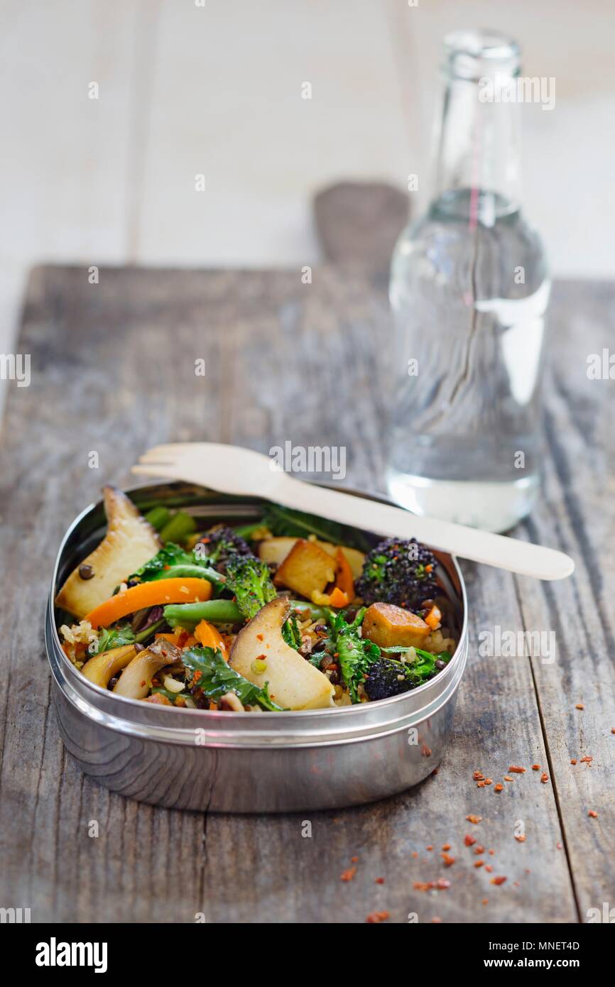 Le lenticchie e il riso con i funghi, le carote e i broccoli in una scatola di pranzo davanti ad una bottiglia di acqua Foto Stock