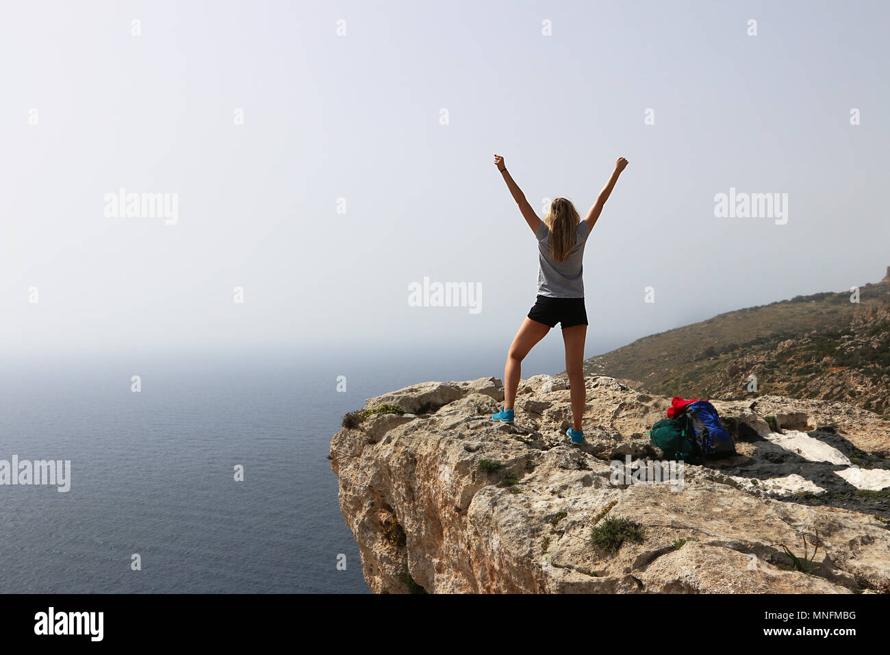 Trionfa il giovane donna a braccia alzate celebra dopo una salita con successo. In piedi torna alla telecamera che guarda al mare dal ciglio della scogliera. Copia dello spazio. Foto Stock